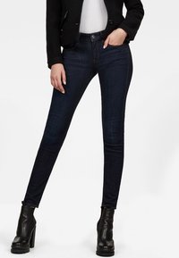 Jeans pitillo de denim azul oscuro, con un diseño clásico de cinco bolsillos y cierre de botón frontal, combinados con botas negras hasta el tobillo.