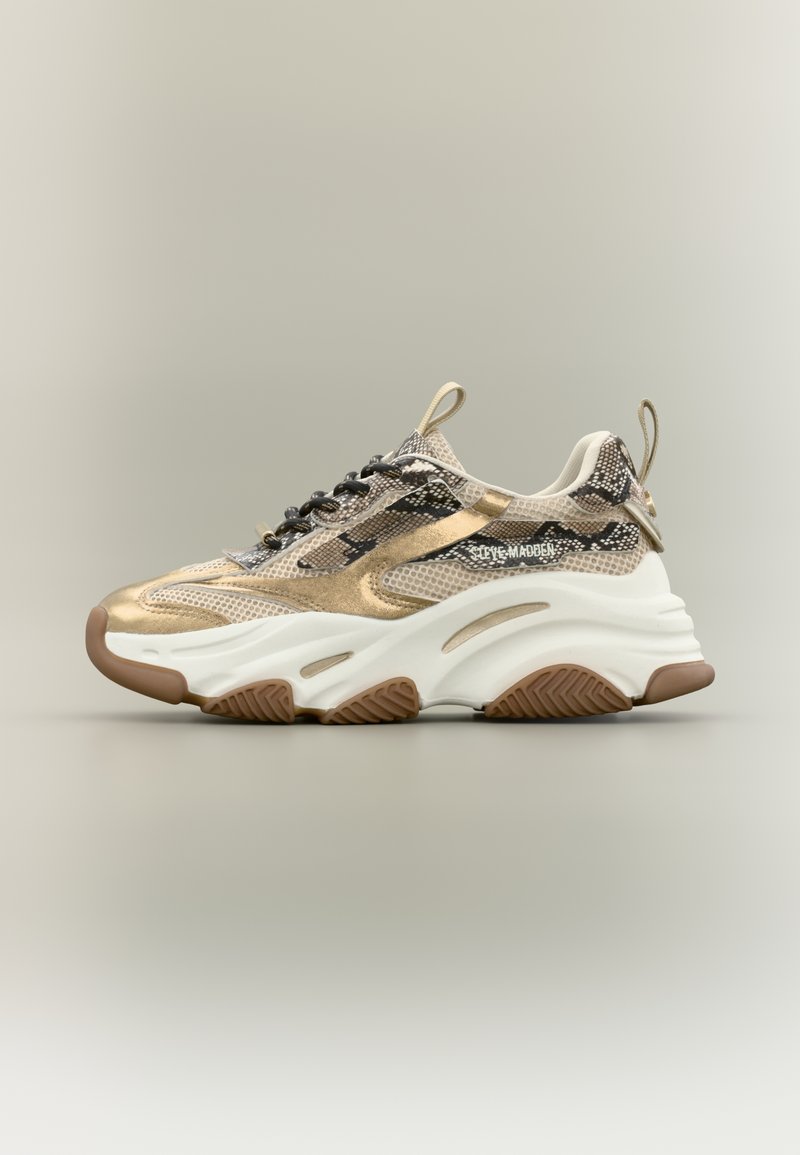 Sneaker met een metallic goud-zwart kleurenschema, bovenwerk van mesh, dikke witte midzool en een gestructureerde bruine rubberen buitenzool.