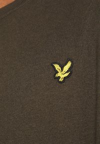 Pull côtelé vert olive avec un col en V et un logo aigle brodé jaune sur le côté gauche de la poitrine. Texture douce, design décontracté.