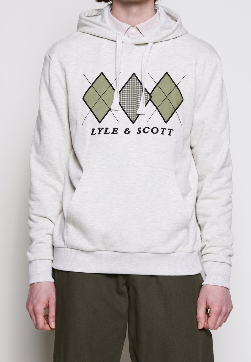 Sweat à capuche gris clair avec motif en losanges verts et à carreaux et texte « LYLE & SCOTT » sur le devant, porté avec un pantalon olive foncé.