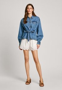 Jeansjacke mit einem taillierten Gürtel, langärmlig und mit gefütterten Brusttaschen. Getragen über weißen, gewellten Shorts und braunen, geschnallten High Heels.