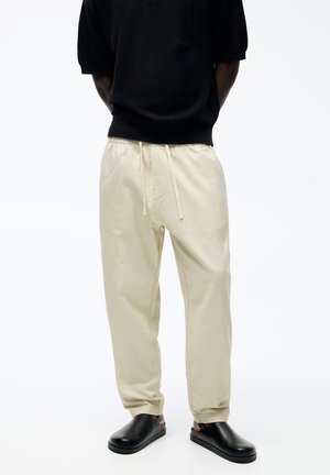 Personne portant un pantalon beige à cordon de serrage, des chaussures noires sans lacets et un pull noir à manches courtes, debout devant un fond blanc uni.