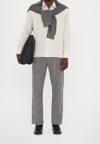 White, striped button-up shirt with a gray knit sweater tied around the shoulders, gray checkered trousers, and glossy black shoes. Black bag in hand.