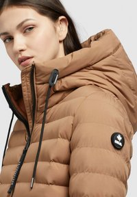 Smeđa puffer jakna s visokim ovratnikom, podesivom kapuljačom i crnim detaljima. Ima romboidni uzorak i okrugli logotip na rukavu.