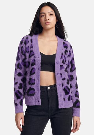 Junge Frau mit langen dunklen Haaren, die eine violette, flauschige Strickjacke mit Leopardenmuster, ein schwarzes Crop-Top und hoch taillierte schwarze Jeans trägt, stehend vor weißem Hintergrund.