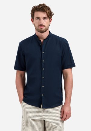 Mann mit braunen Haaren, der ein marineblaues kurzärmeliges Hemd mit Knöpfen und beigefarbene Hosen mit schwarzen vertikalen Streifen trägt, die Hände in den Taschen.