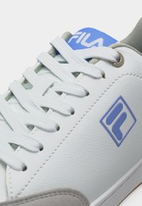 Κοντινό πλάνο σε λευκό αθλητικό παπούτσι Fila με λευκά κορδόνια, μπλε λογότυπο στο πλάι και λεπτομέρεια με μπλε σήμανση στη γλώσσα.