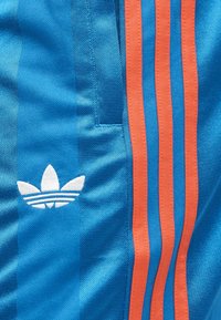 Μπλε ύφασμα με κόκκινες κάθετες ρίγες και λευκό λογότυπο τριφυλλιού, εμφανίζοντας υφή ραψίματος και ελαφρές πτυχές υφάσματος.