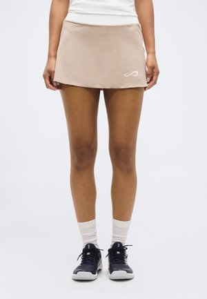 Woman wearing beige sports skort, white socks, and black athletic shoes, standing against a plain light background.