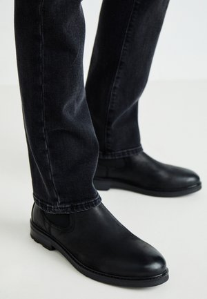 Botas de tobillo de cuero negro con punta redonda y paneles laterales elásticos, llevadas con jeans de mezclilla oscuros, mostrando una textura suave y un diseño aerodinámico.