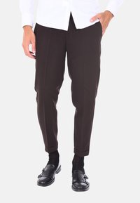 Pantaloni marroni su misura con una vestibilità affusolata, caratterizzati da una texture liscia, linee di piegatura e risvolti sopra le caviglie. Indossati con mocassini neri.