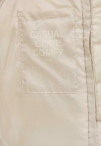 Beigefarbene Stoffetikette, die auf dem Kleidungsstück genäht ist, mit dem Text "LÄSSIG COOL BEQUEM", Markenname sowie Symbolen für recyceltes Polyester und Pflegehinweise.