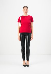 Woman wearing a red short-sleeve top, black leather pants, black heels, and carrying a white shoulder bag, standing against a plain white background.