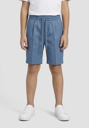 Kind draagt blauwe shorts met trekkoord, witte sneakers en een wit T-shirt, staand voor een effen grijze achtergrond.