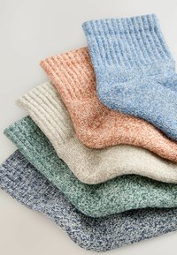 Pile de cinq chaussettes en maille texturée de couleur bleue, orange, beige, verte et marine, disposées sur une surface claire.