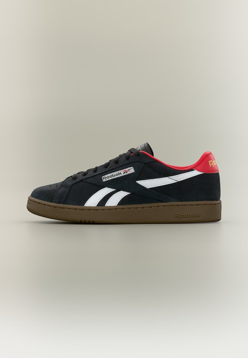 Zapatilla negra de Reebok con franjas laterales blancas, collarín rojo en el talón, suela marrón y cordones negros, mostrada de perfil sobre un fondo neutro.