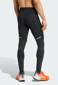 Black athletic leggings featuring a smooth finish, mesh side panels, reflective accents, and a zippered pocket. Paired with orange and gray shoes.