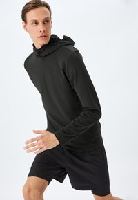 Sort langærmet atletisk hoodie med høj krave og afslappet pasform. Parret med sorte shorts, der har en glat, strækbar tekstur.