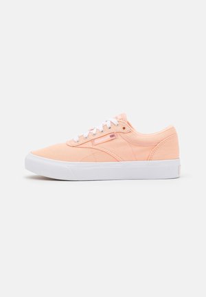 Zapatilla de lona de color melocotón claro, tipo baja, con cordones y suela blancas, con el logo de Reebok y detalle de la bandera británica en el lateral.