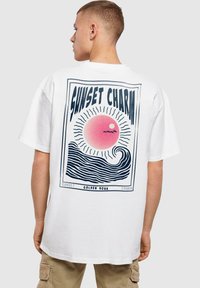 Camiseta de algodón blanca con un gráfico de estilo retro que incluye un sol rosa y olas estilizadas. El texto dice "ENCANTO DEL ATARDECER" en una fuente en negrita.