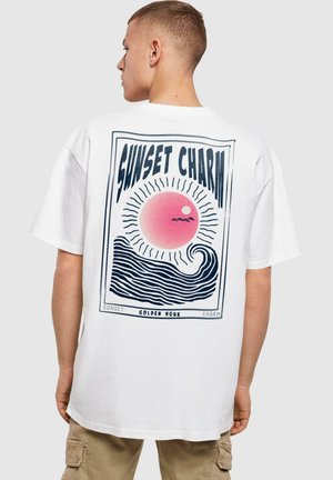 Hvit bomulls t-skjorte med et retro-inspirert grafisk design med en rosa sol og stiliserte bølger. Teksten sier "SUNSET CHARM" i fet skrift.