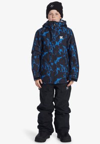 Svart och blåmönstrad snöjacka med huva, bärs med lösa svarta snöbyxor och svarta snöskor. Har logotypdetalj på jackan.