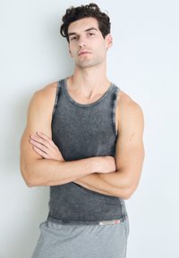Grå tanktopp med en falmet, vasket tekstur og rund hals, sammen med grå shorts, modellen har armene krysset mot en lys bakgrunn.