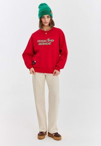 Czerwona bluza z zielonym napisem "mucho amor", luźny krój, zestawiona z jasnobeżowymi spodniami i brązowymi butami. Na głowie zielona dziana czapka.