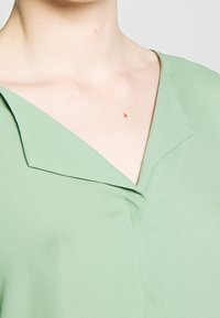 Blusa verde con scollo a V e design del colletto angolare. Tessuto liscio, leggero e con dettagli di cucitura sottili.