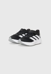 Chaussures de sport noires avec des rayures blanches, tige en mesh, bout arrondi, sangle ajustable et semelle extérieure grise. Design minimaliste.