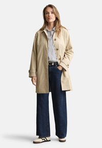 Gabardina beige con grandes botones, llevada sobre una camisa a rayas, combinada con vaqueros de pierna ancha en denim oscuro y zapatillas beige con detalles negros.