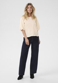 Blouse en crème texturée avec des manches bouffantes, associée à un jean en denim foncé taille haute et des bottines noires. Design simple et décontracté.