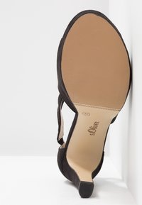 Svart mocka högklackad sko med spetsig tå, smal klack och strukturerad sula. Har en präglad logotyp på innersulan.