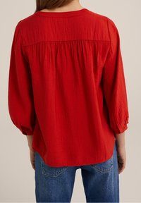Red woven blouse with a pleated back, three-quarter sleeves, and a rounded neckline, paired with blue denim jeans. Textured fabric.