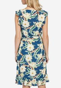 Kvinna som bär en ärmlös blå klänning med gult och vitt blommigt mönster, volangkant och ett midjeband, sedd bakifrån.