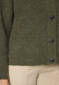 Olivgrön, texturerad stickad kofta med rund halsringning, sex svarta knappar och ribbade ärmslut, tillsammans med ljusa byxor.