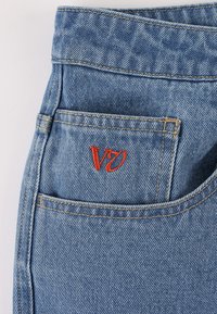 Lyseblå denimjeans med synlig tekstur. Har en baglomme med rødt broderet "V7"-logo og kontrastfuld gul syning.