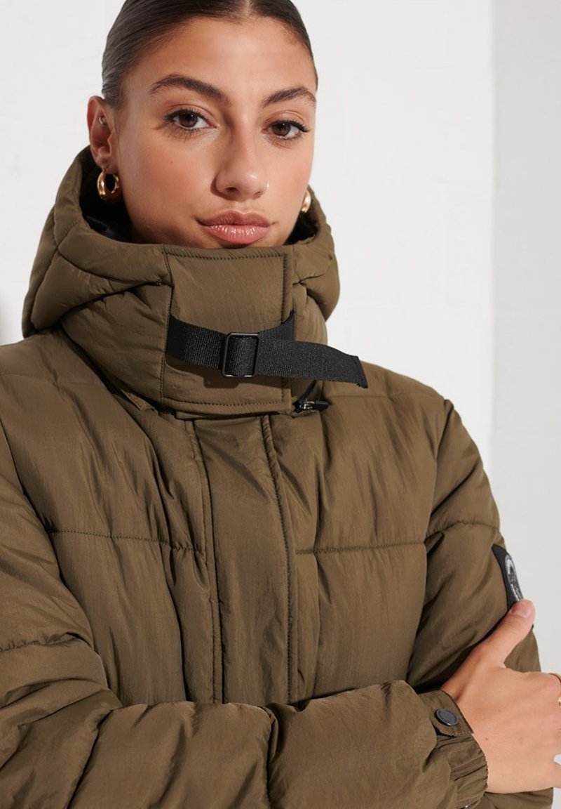 Mujer con el cabello recogido, que lleva pendientes de aro dorados y una chaqueta acolchada verde oliva de cuello alto con una correa negra de hebilla.