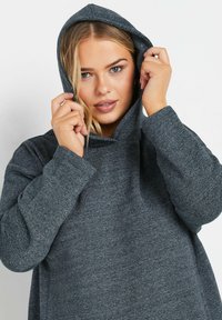 Sudadera con capucha gris hecha de una tela suave, con un corte holgado y mangas largas. La capucha está ajustada alrededor de la cara.