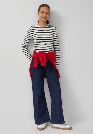 Gestreept langemouwtop met uitsneden op de schouders, gecombineerd met een brede, donkerdenim spijkerbroek en een knopenrode sweatshirt om de taille. Witte sneakers.