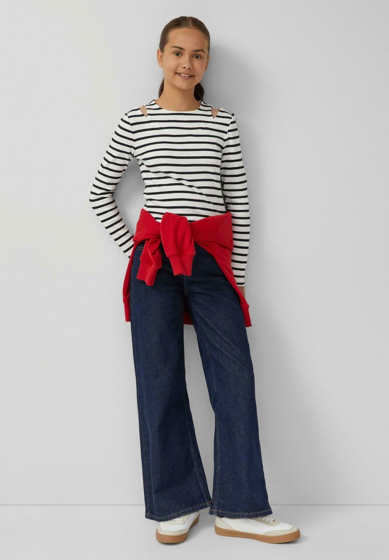 Gestreiftes Langarmoberteil mit Cut-Out-Schulterdetails, kombiniert mit weiten dunklen Jeans und einem gebundenen roten Sweatshirt in der Taille. Weiße Sneaker.