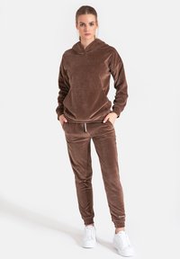 Brązowa bluza z weluru z kieszenią z przodu i pasującymi dresami. Dopasowany krój z elastycznymi mankietami; zestawiona z białymi trampkami. Gładka faktura.