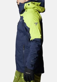 Giacca impermeabile blu navy e verde lime con cappuccio, multiple tasche con cerniera e polsini regolabili. Presenta il marchio "Omni-Tech" sulla manica.