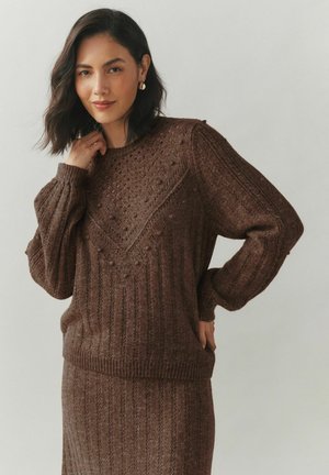 Frau mit schulterlangem dunklem Haar, die einen braunen, strukturierten Strickpullover und einen passenden Rock trägt und mit einer Hand nahe am Hals posiert.