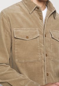 Camisa de pana en beige, con cierre delantero de botones, dos bolsillos en el pecho con solapas y cuello clásico. Tejido acanalado con textura.