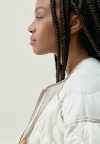 Nafoukaná bílá bombera s předním zipem, texturovaná tkanina a béžové akcenty. Model má dlouhé, spletené vlasy.