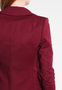 Blazer ajustado burdeos con una textura suave, cuello de muesca y sutilezas en la costura, que presenta una silueta entallada y un diseño elegante.