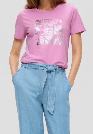 Frau trägt ein pinkes T-Shirt mit silbernem floralem Quadratmuster und hellblaue hoch taillierte Jeans, die in der Taille gebunden sind.