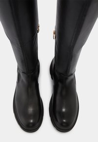 Bottes en cuir noir au-dessus du genou avec un design épuré, une texture lisse et un détail en métal doré pour la fermeture éclair. Semelle noire épaisse.