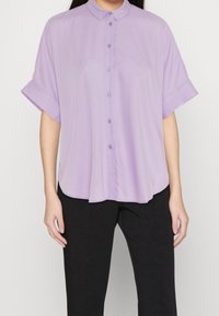 Světle fialová košile s krátkým rukávem, zapínáním na knoflíky a límečkem, měkkou texturou, zaobleným lemem a ohrnutými rukávy. Spárována s černými kalhotami.
